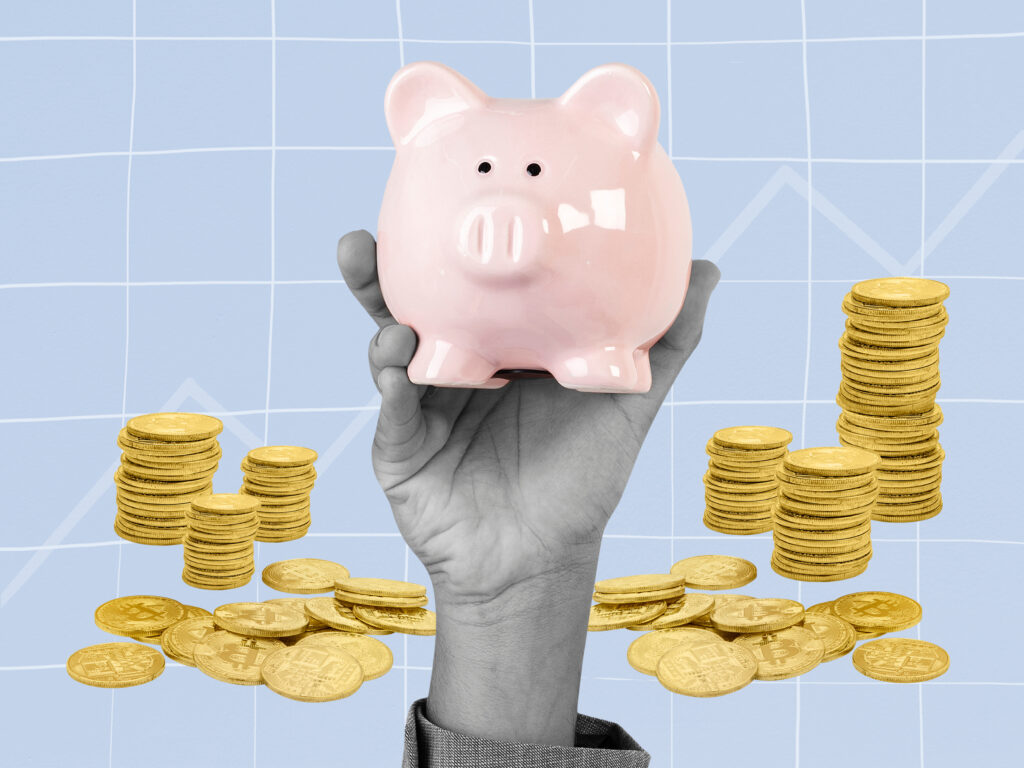 A hand is holding a piggy bank with stacks of coins in the background.