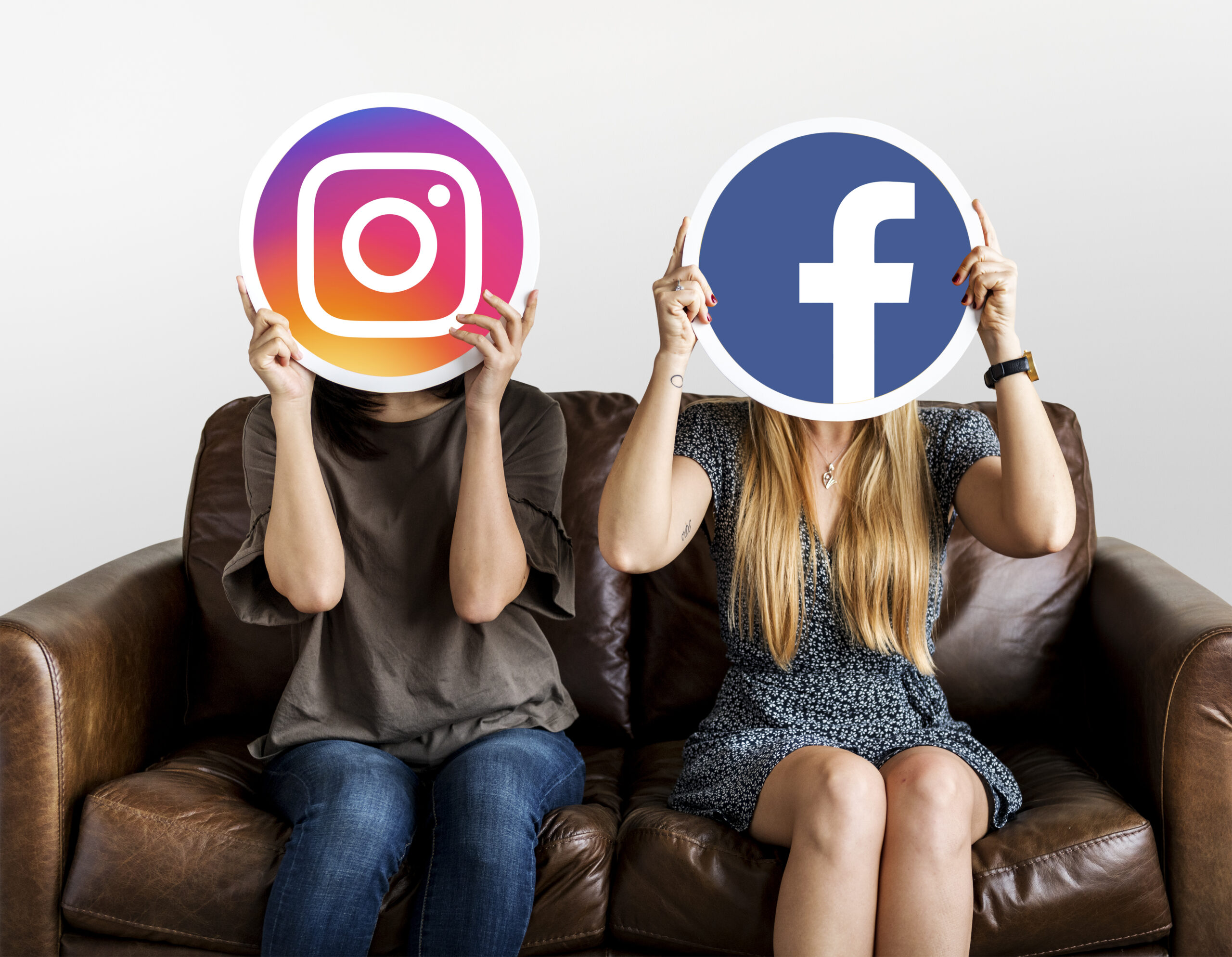 Two women are seated on a couch. One is holding the Instagram logo in front of her face, the other is holding the facebook logo in front of hers.