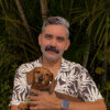 An Aboriginal man with a mustache and a tropical shirt is holding a brown sausage dog.