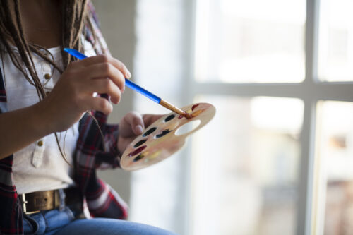 A young women is holding a palette and paintbrush with a light filled window behind her.