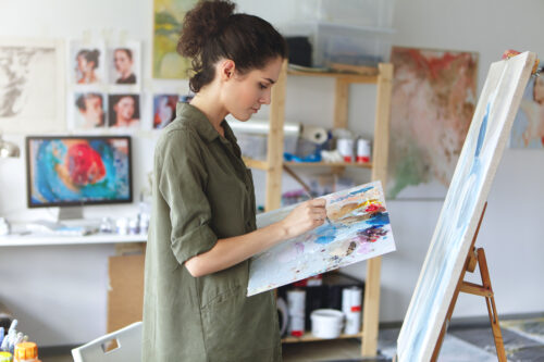 Student girl of faculty of arts wearing military green t-shirt standing in front of easel, mixing colors using paint brush while working on painting in university studio. Inspiration and creativity