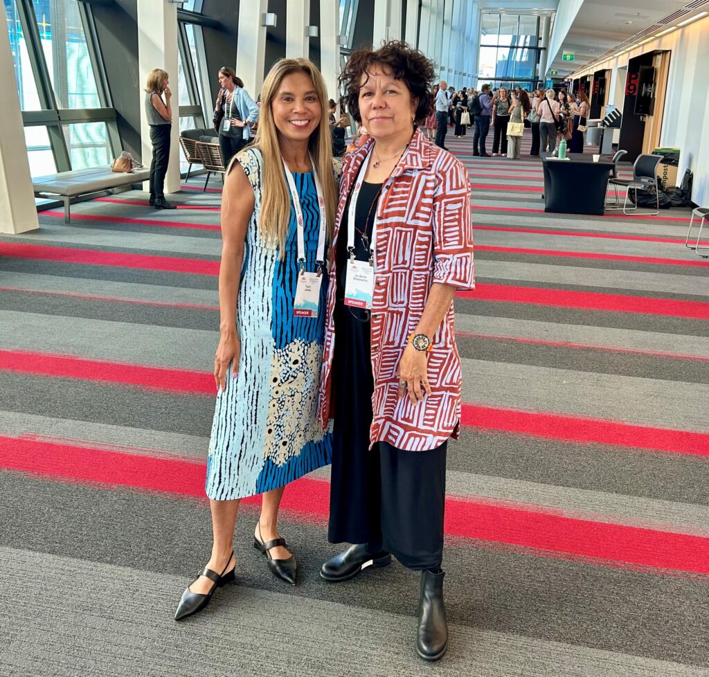 Two women weraring brightly coloured Aboriginal printed dresses standing in a large conference room.