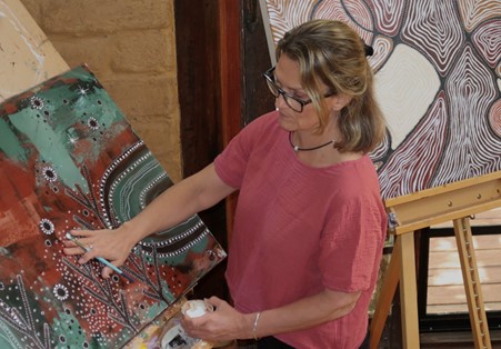 A blonde woman in a pink tshirt is working on a large colourful painting.