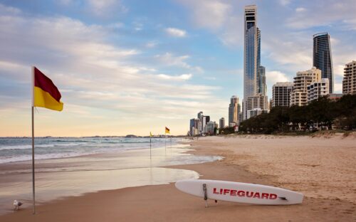 A beach with a red and yellow flag and a surf lifesaving board. There are skyscrapers in the background.